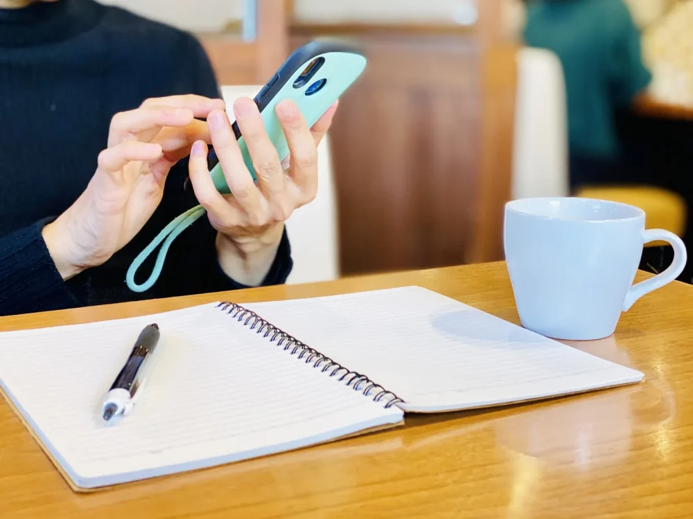 ノートとコーヒーが置かれた机でスマートフォンを操作する手元